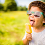 The tongue of a young boy with curly hair is touching an orange frozen treat. He is wearing a gray t-shirt, a straw hat and blue rimmed glasses. The background is blurry and consists of an open field of green grass with trees and bushes in the distance.