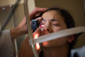 An ophthalmologist shines light into the eye of a tense young female patient who has a piece of foreign object (metal shard) stuck in her cornea. Real injury and examination. A slit lamp instrument for closer examination is in the foreground and lights the doctor’s hand.