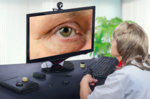 Telemedicine female ophthalmologist looks at epidermal cyst on right upper eyelid on monitor seriously. Virtual doctor observes mans cyst either by online video conference or snapshot. Horizontal mid-shot on blurry indoors background