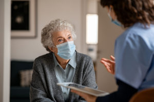 Senior woman wearing safety protective mask at home and talking to nurse holding digital tablet. Back view of young doctor visiting old woman for routine health checkup during covid-19 pandemic. Young general practitioner and elderly patient wearing face masks in a private medical consult during coronavirus and flu outbreak.
